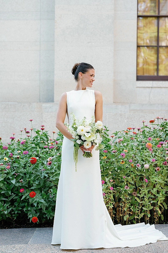 Bridal portrait of a bride in a minimalist wedding dress holding a white and green calla lily bouquet beside a stone wall and garden greenery