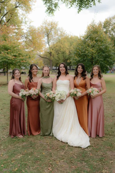 Bride and bridesmaids in mismatched bridesmaid dresses gather beneath autumn park trees, holding blush and ivory bouquets on the lawn.