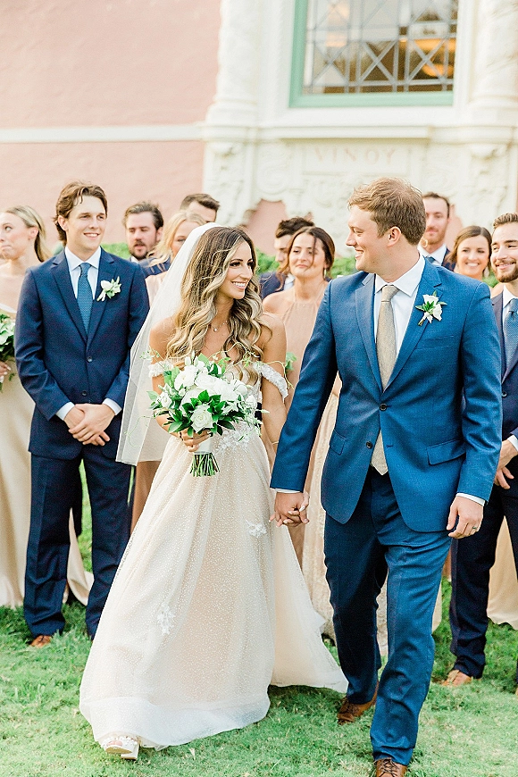 Wedding recessional as bride and groom walking hand in hand, bouquet and veil flowing, wedding party behind on a grass lawn by windows