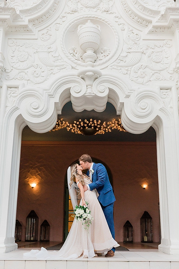 Wedding kiss portrait of the bride and groom kissing in a dip, her cathedral veil flowing as they stand beneath an ornate white archway.