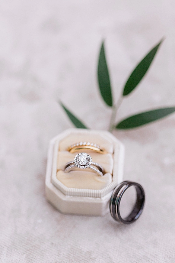 Wedding rings and engagement ring close up on a ring box with a greenery sprig, set on white textured fabric backdrop