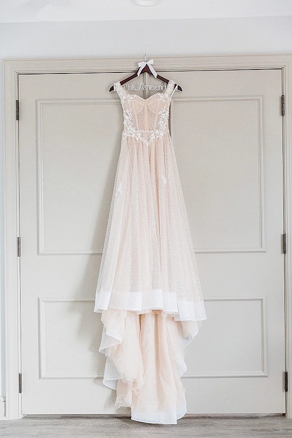 Wedding dress with lace bodice and blush tulle skirt hanging from a personalized wire hanger on double doors indoors