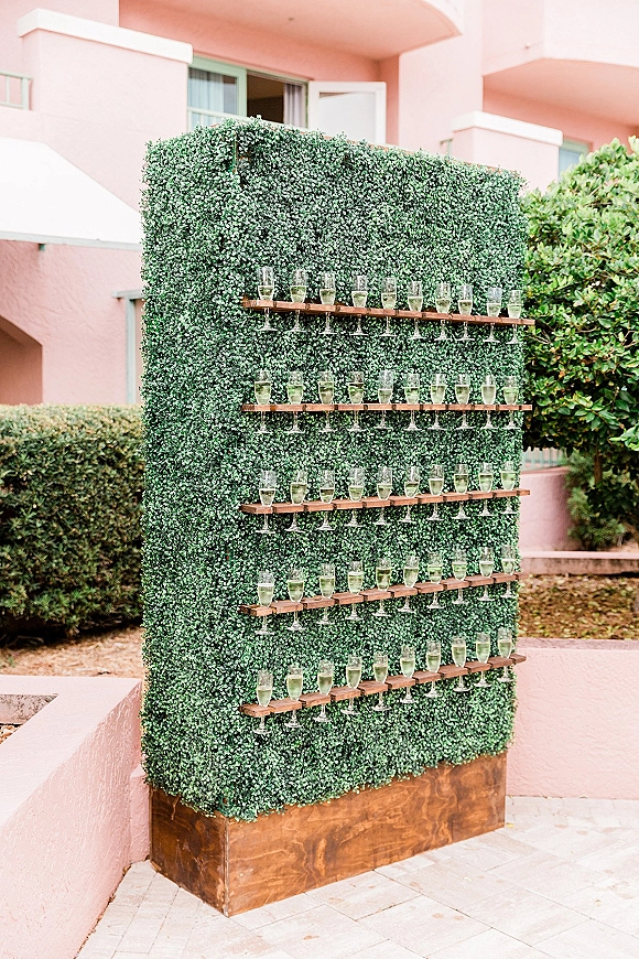 Champagne wall with wooden shelves of pre-poured flutes against a greenery hedge, set on patio pavers by a pink exterior courtyard