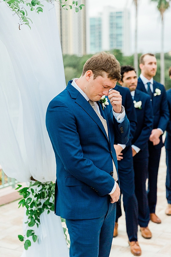 Groom crying, wiping tears in a navy suit with boutonniere beside groomsmen at an outdoor ceremony with draped greenery arch