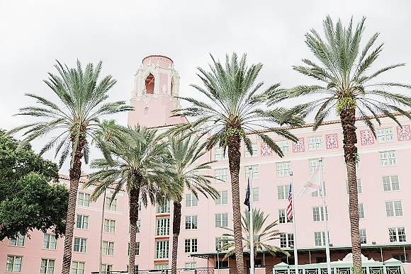 Wedding venue exterior of a pink hotel wedding venue with palm trees, flags, arched windows and tower under an overcast sky