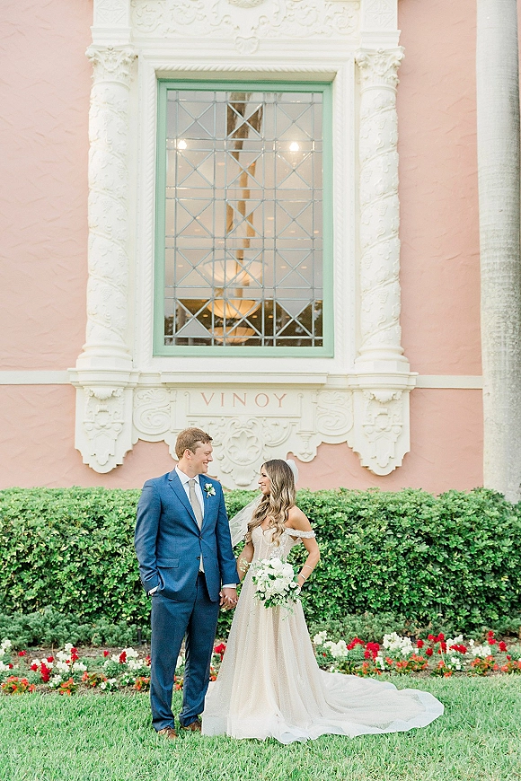 Couple portrait of bride and groom holding hands, bride with white bouquet and veil, beside a pastel pink wall with ornate window backdrop