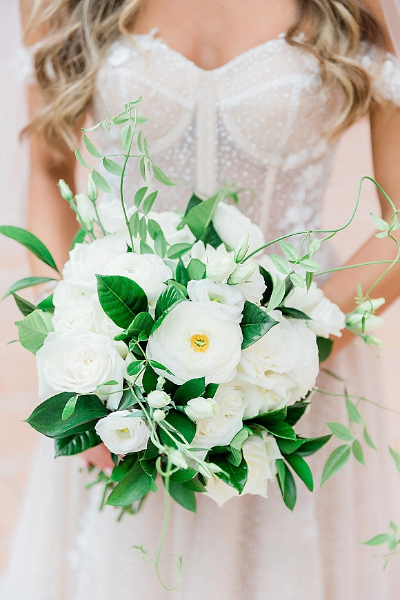 Bridal bouquet of white wedding bouquet blooms with lush greenery, held against an ivory bridal gown bodice on a neutral backdrop