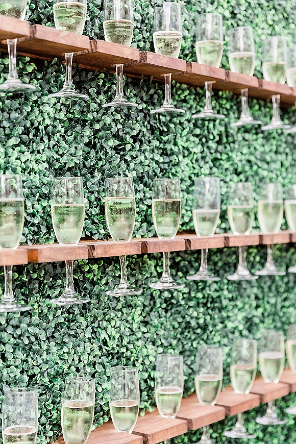 Champagne wall with wedding champagne wall shelving, champagne flutes and bottles on wooden shelves against a lush greenery backdrop