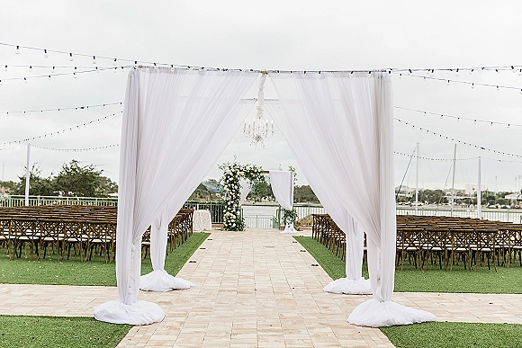 Ceremony aisle design with sheer draping and chandelier over a white runner, leading to a floral arch on a waterfront lawn with sailboats