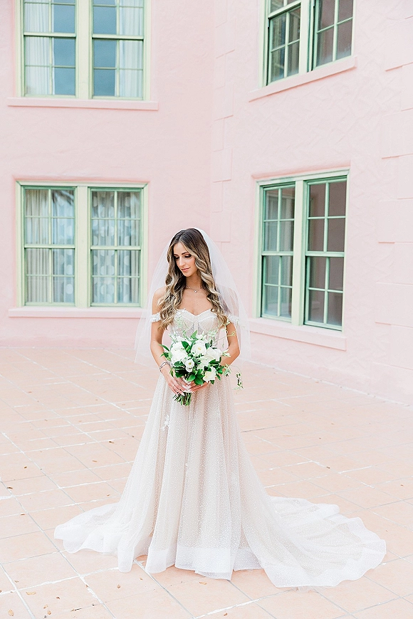 Bridal portrait of a bride holding bouquet in a strapless gown and veil, standing by a pink stucco wall with green windows on a tiled patio