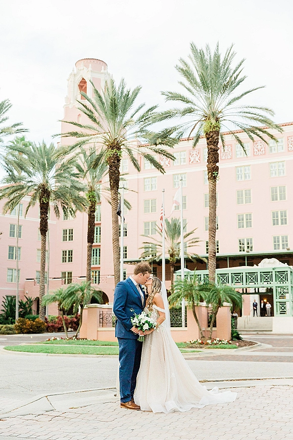 Wedding kiss portrait of bride and groom kissing, her white veil and bouquet in hand, framed by palm trees and a hotel driveway backdrop