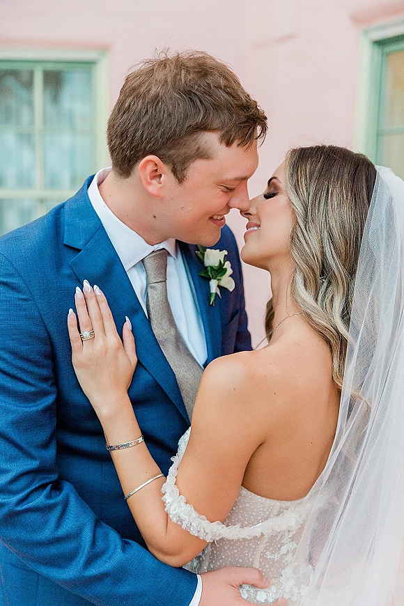 Wedding couple portrait of bride and groom close up touching foreheads, bride showing ring, veil and lace dress against a pink wall by a window
