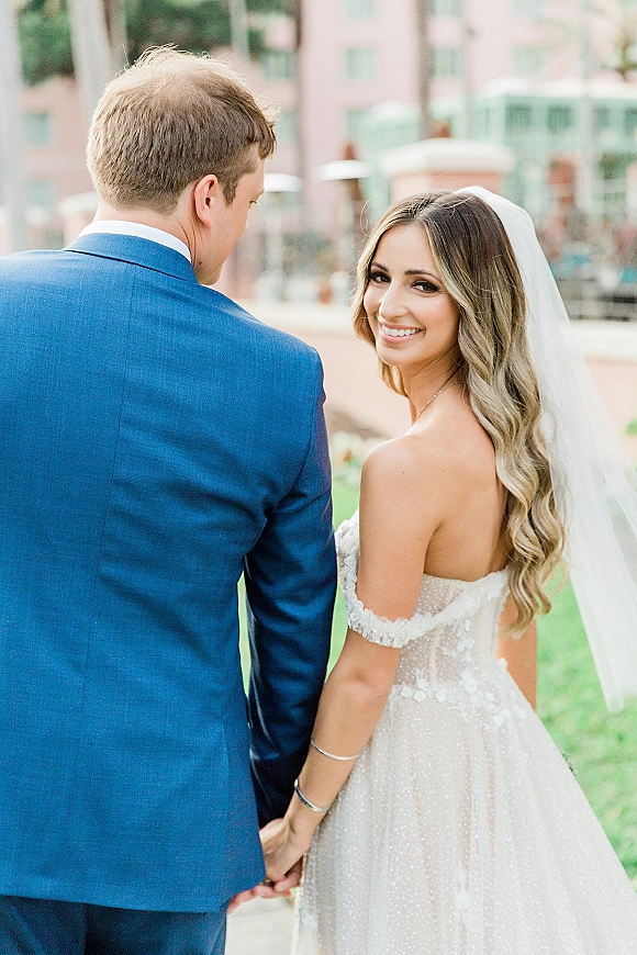 Couple portrait of bride looking back while holding hands with groom in blue suit, walking on a lawn by a modern building with veil flowing