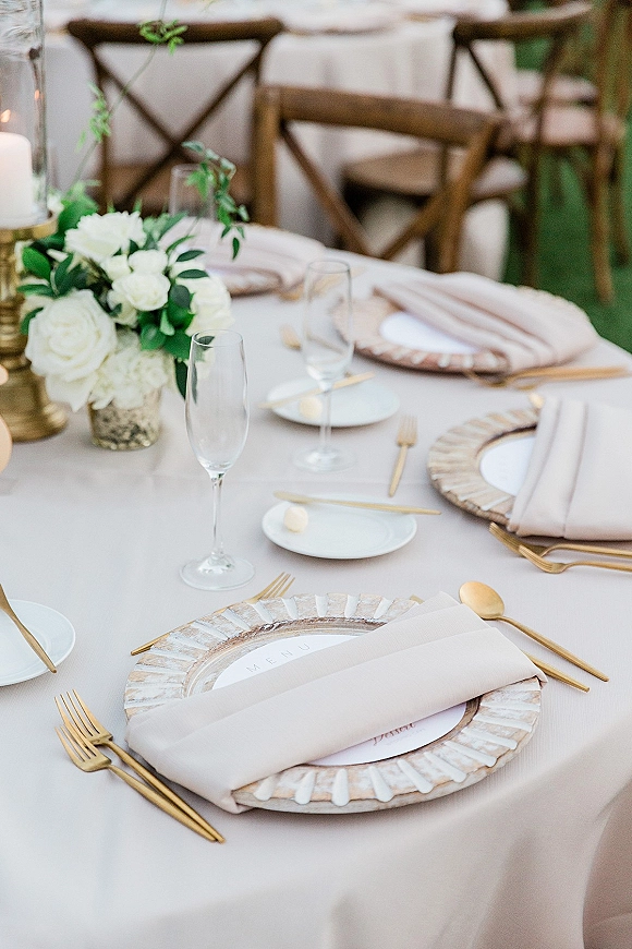 Reception tablescape with a wedding table setting featuring blush napkins, gold flatware, white florals, taper candles, and crossback chairs outdoors