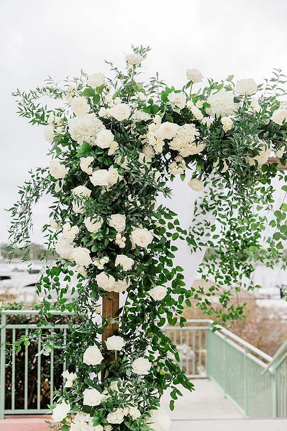 Wedding floral arch with white rose clusters and trailing greenery garland on a wooden frame along an outdoor walkway with trees and sky