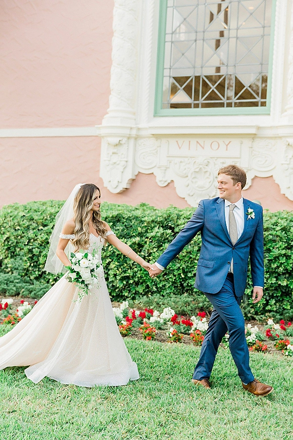 Couple portrait of bride and groom walking hand in hand, her veil flowing and bouquet held, beside hedges and a pink facade