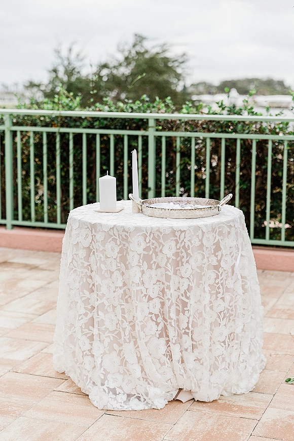 Unity candle table with a wedding unity candle set on a silver tray atop a lace tablecloth on an outdoor patio with greenery behind