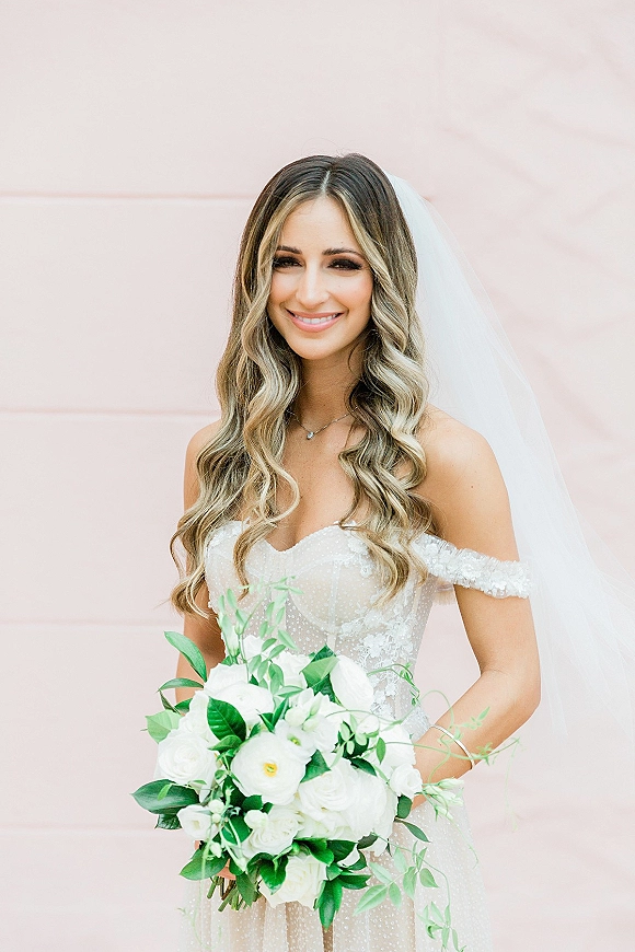 Bridal portrait of a smiling bride holding bouquet of white roses with greenery, wearing an off-the-shoulder lace dress before a pink wall