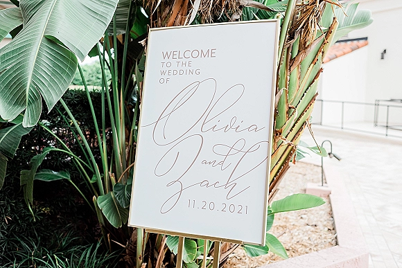 Wedding welcome sign in a gold frame on an easel with modern calligraphy and palm leaf accents along an outdoor walkway by greenery