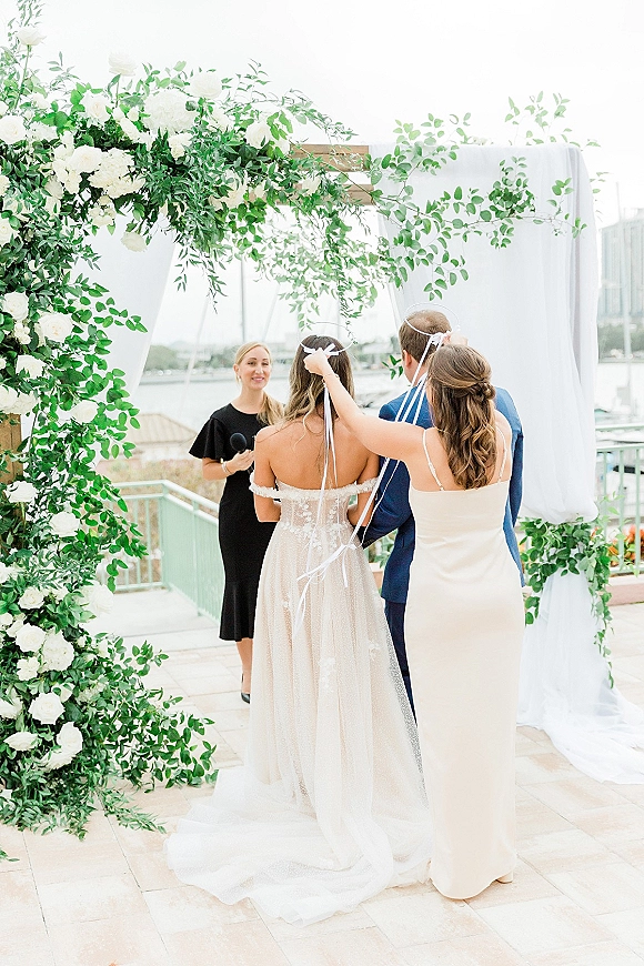 Wedding ceremony moment with bride and groom at altar under a white rose and greenery arch, waterfront marina and boats behind under cloudy sky