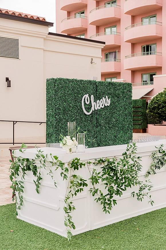 Wedding bar setup with a wedding bar backdrop of boxwood hedge, white counter, greenery garland, glassware, and cocktail sign on a hotel patio lawn