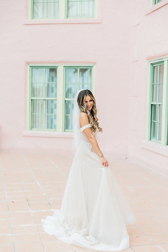 Bridal portrait of a smiling bride in an off the shoulder wedding dress with a long veil, looking back by pink wall and mint windows