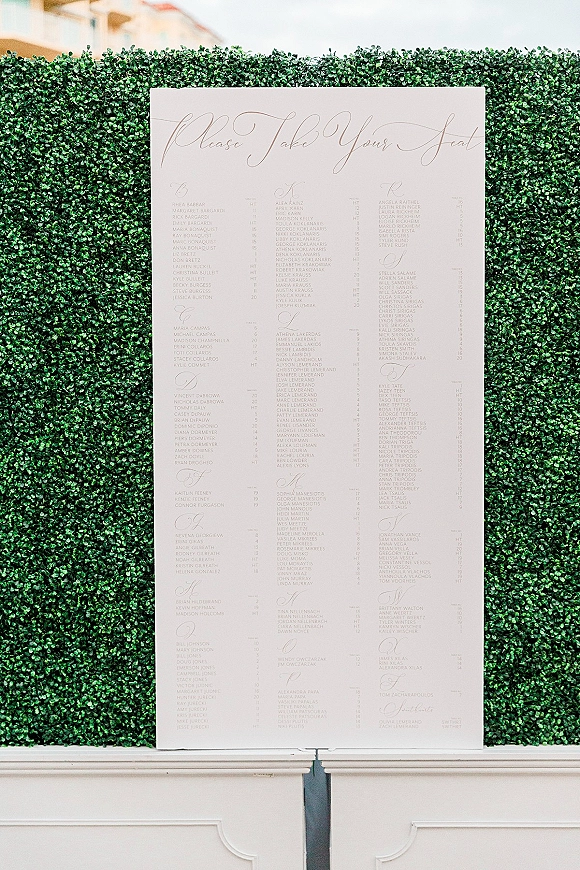 Wedding seating chart with calligraphy seating chart lettering on a white board displayed on an easel against a boxwood hedge wall and wainscoting