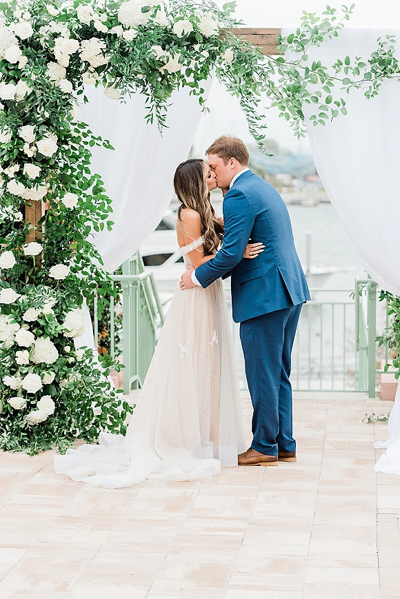 Wedding kiss portrait of bride and groom kissing under arch with white roses and drapery on a waterfront patio with boats behind