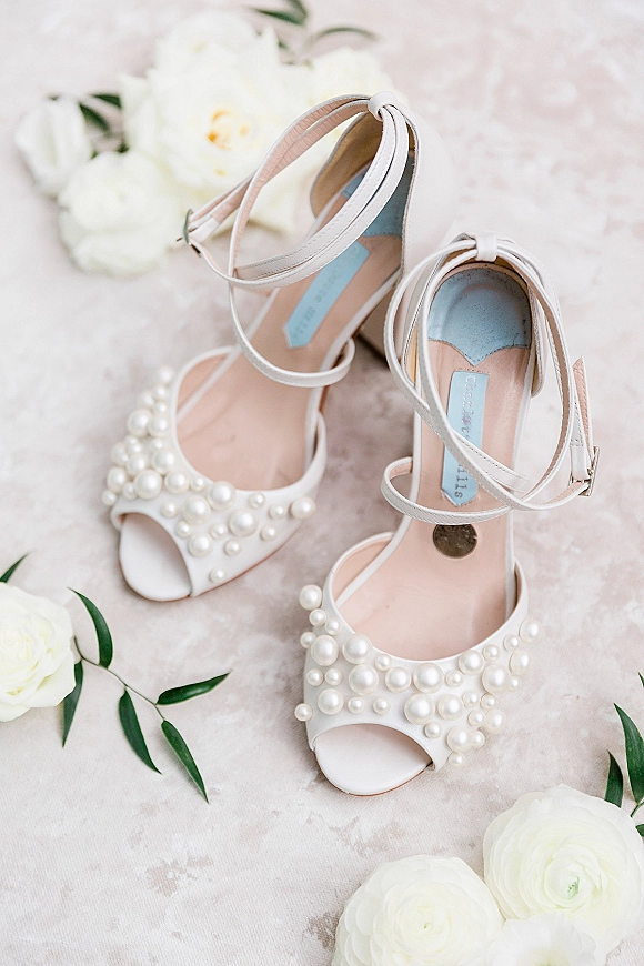 Wedding shoes with pearl wedding heels and ankle straps in a flat lay with white flowers and greenery on a blush textured surface
