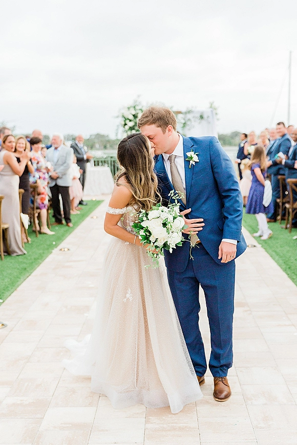 Wedding kiss as bride in an off-the-shoulder dress holds a white rose bouquet, groom in blue suit on aisle runner with guests outdoors