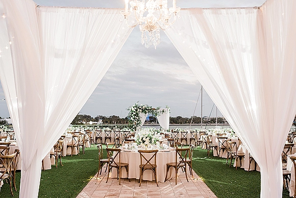 Outdoor wedding reception under a white draped tent with crystal chandelier and string lights, set with round tables on a waterfront lawn