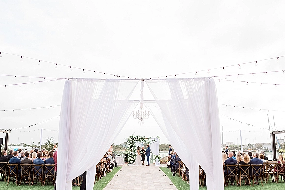 Wedding ceremony under a white draped canopy with chandelier, floral arch and aisle runner on an outdoor terrace with water view