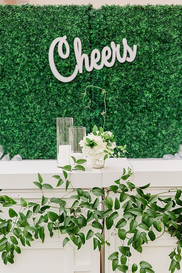 Wedding bar backdrop with boxwood hedge backdrop, a Cheers sign, white roses and greenery garland above a white bar counter