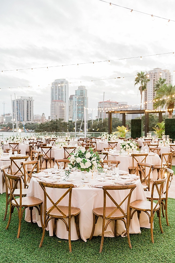 Outdoor reception setup for a rooftop wedding reception with round tables, ivory linens, cross back chairs, and string lights by the city skyline