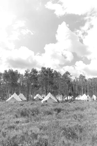 Glamping wedding tents with canvas accents arranged across meadow grass near forest trees under a cloudy sky for guest lodging
