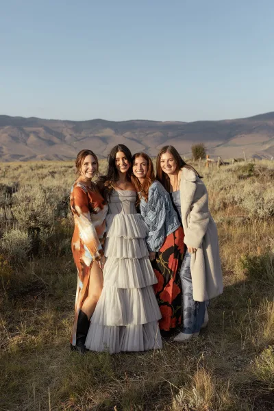 Bridesmaid group photo with mix and match dresses, one in faux fur coat, standing in a dry field with mountains and blue sky behind