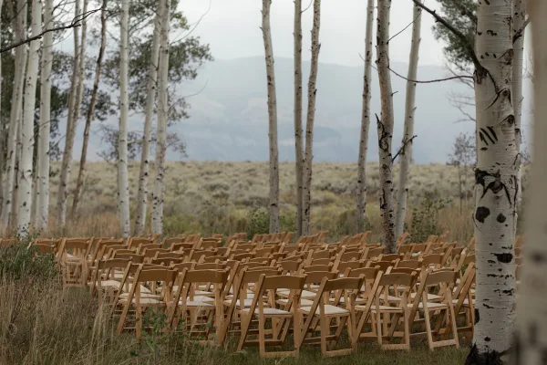 Outdoor ceremony setup with wood folding chairs and cushions in neat rows, set in an aspen meadow with mountains under an overcast sky
