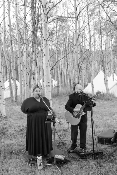 Wedding musicians performing live music with acoustic guitar and microphones in an aspen forest near white tents at an outdoor ceremony