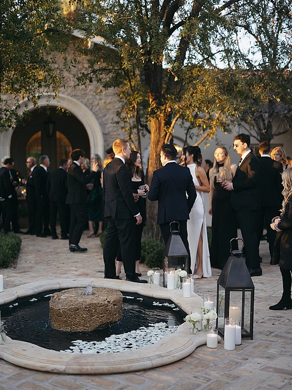 Wedding cocktail hour with guests in formalwear mingling around a courtyard fountain, cocktail glasses in hand, lanterns glowing nearby