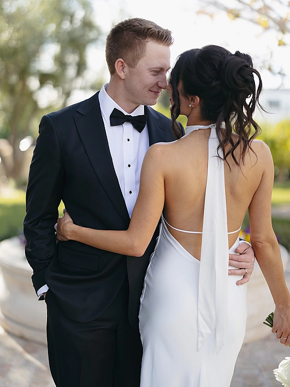 Couple portrait of bride and groom embrace, forehead touching; she wears an open-back gown with bouquet beside outdoor greenery walkway.