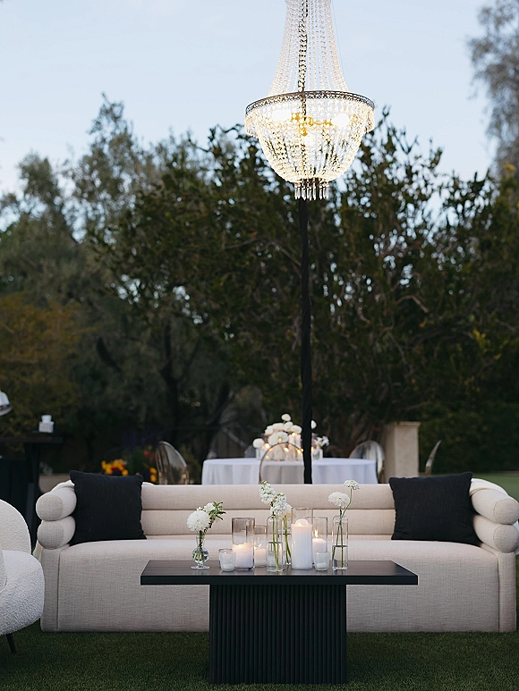 Wedding lounge seating with a crystal chandelier above a sofa, candles and bud vases on a coffee table, set on a garden lawn near dining tables
