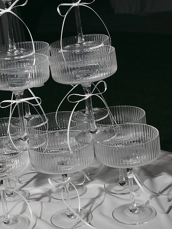 Champagne tower of coupe glasses with white ribbon bows stacked on a table, styled as a wedding champagne display against a dark backdrop