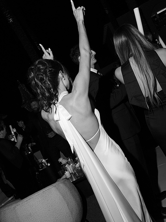 Wedding reception dancing as the bride raises her arms in a backless dress with bow detail, surrounded by guests and taper candles in a dim lounge room