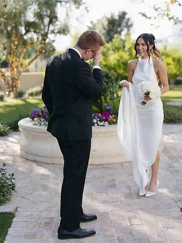 Wedding first look as bride in sleek halter dress and veil holds white rose bouquet, facing groom in black tux under string lights in a garden