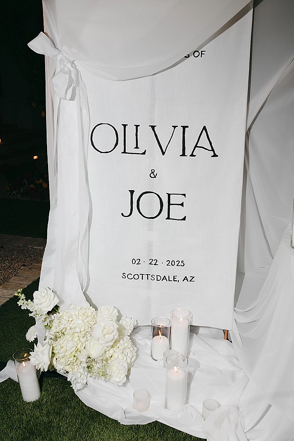 Wedding welcome sign with personalized names, draped fabric, white roses and hydrangea, and candle clusters on a lawn at night