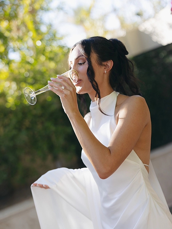 Bride portrait sipping champagne from a flute, showing her engagement ring and halter open-back white dress in sunlit greenery bokeh