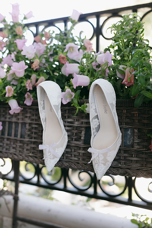Bridal shoes, white lace wedding heels with bow detail nestled in a wicker basket among pink bell flowers on a bright balcony railing