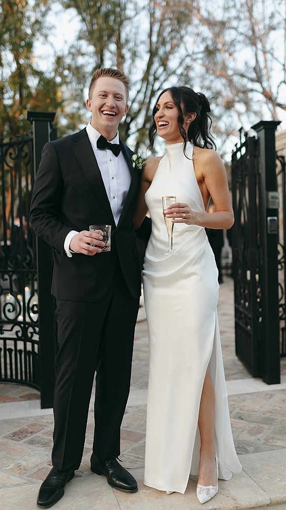 Couple portrait of bride and groom laughing, holding champagne and whiskey in front of a wrought iron gate on a stone patio