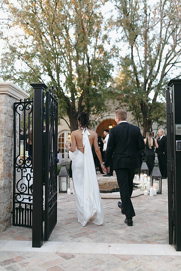 Couple walking hand in hand, bride and groom walking away in a stone courtyard past lanterns and pillar candles toward an arched doorway