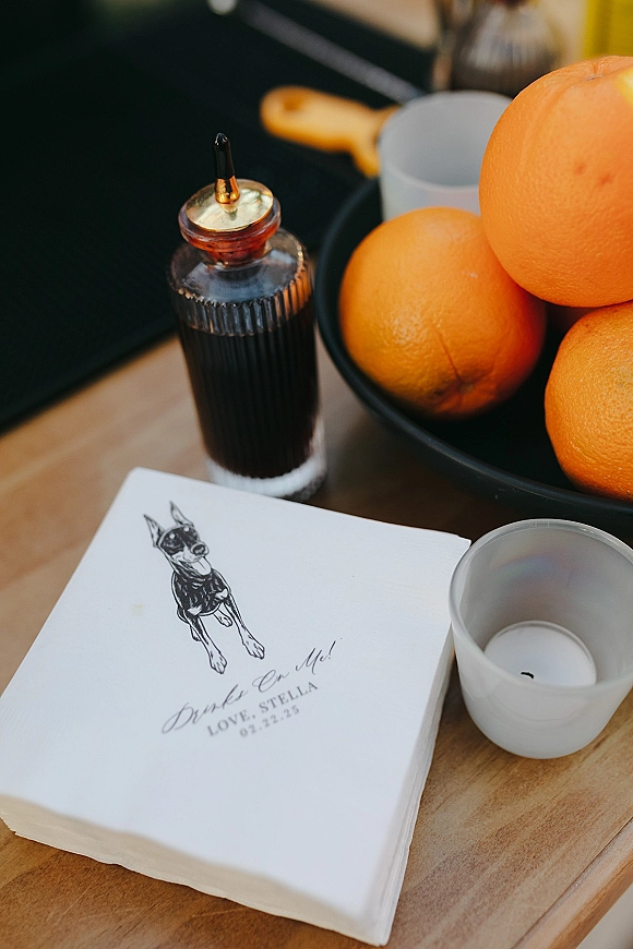 Wedding cocktail napkins, custom wedding napkins with an illustrated dog, sit beside oranges and a bitters bottle on a wood bar counter